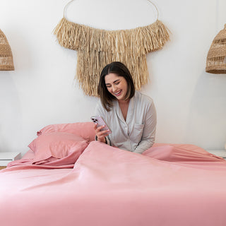 lady sitting in bed under sunset blush bamboo sheets laughing whilst looking at her phone