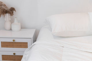 a white bamboo quilt with white sheets and a white pillowcase on a mattress with a white and wood sideboard